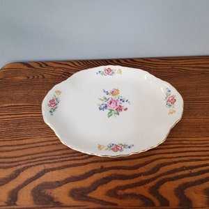 Puede incluir: Plato ovalado blanco con borde festoneado y ribete dorado. El plato está decorado con racimos de flores rosas, rojas, amarillas y azules. El plato está sobre una mesa de madera.