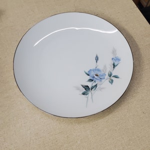 Puede incluir: Un plato de cena de cerámica blanca con un borde plateado presenta un diseño floral azul con una sola flor grande y dos botones más pequeños.