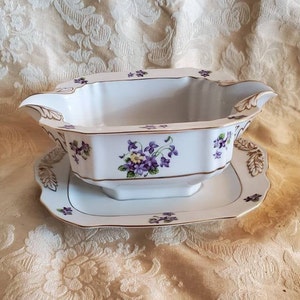 Puede incluir: Salsera de porcelana blanca con ribete dorado y diseño floral morado y amarillo. La salsera está sobre un plato a juego.