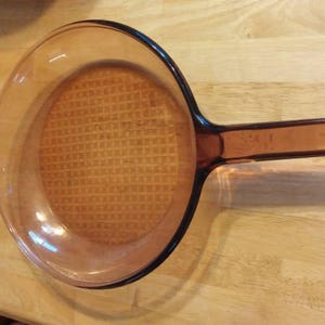 May include: A brown glass frying pan with a waffle-like texture on the bottom. The pan has a brown handle.