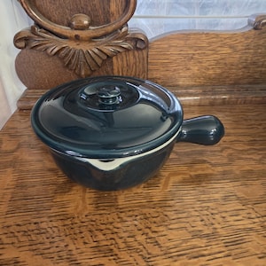 Op de afbeelding: Een donkerblauwe keramische soepkom met een bijpassend deksel en een enkele handgreep. De kom heeft een witte rand en een kleine knop op het deksel. De kom staat op een houten oppervlak.