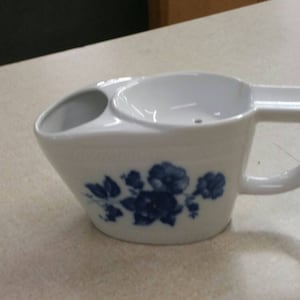 Könnte beinhalten: Weiße Keramik-Rasiertasse mit blauem Blumendesign. Die Tasse hat einen Henkel und einen kleinen Ausguss.