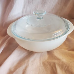 May include: White glass baking dish with a clear glass lid. The dish has a rounded shape and a smooth, white finish. The lid has a knob on top.