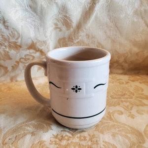 Könnte beinhalten: Weiße Keramiktasse mit einem blauen und weißen Blumenmuster. Die Tasse hat einen Henkel und einen schwarzen Rand.