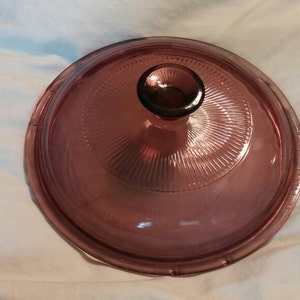 Op de afbeelding: Een paarse glazen deksel met een knop aan de bovenkant. De deksel heeft een getextureerd, geribbeld ontwerp.