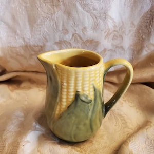 May include: A ceramic jug with a corn on the cob design. The jug is yellow inside and has a handle. The exterior features a detailed depiction of corn, with green husks and yellow kernels. The jug is likely used for serving.