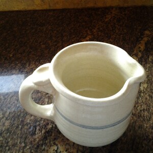 May include: Cream-colored ceramic pitcher with a rounded body and a spout. The pitcher has a handle with a decorative element and a thin blue stripe around the middle. The interior is a lighter cream color.