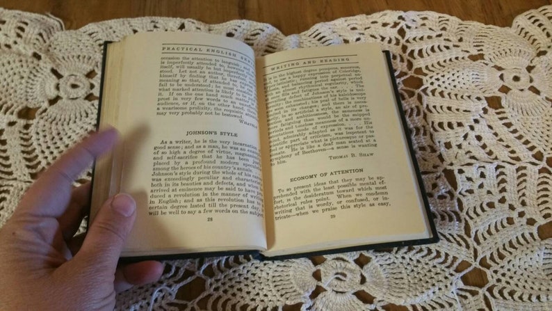 May include: An open book with a black cover and cream-colored pages. The book is titled "Practical English" and "Writing and Reading". The text on the pages discusses the writing styles of Samuel Johnson and Thomas H.  Baw.