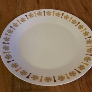 May include: A white ceramic dinner plate with a brown, yellow, and orange floral pattern around the rim.