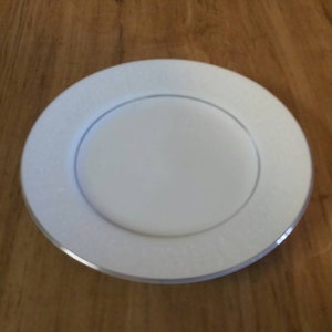 Puede incluir: Un plato de servicio blanco con un borde plateado. El plato es redondo y tiene un borde ligeramente elevado.