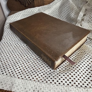 Può includere: Un libro marrone rilegato in pelle con un segnalibro a nastro bordeaux. Il libro è appoggiato su una tovaglietta di pizzo bianco all'uncinetto. Il dorso del libro ha lettere dorate. Lo sfondo include una sedia di legno.