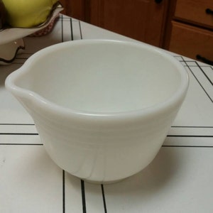 May include: A white ceramic mixing bowl with a pouring spout. The bowl is on a white and black checkered surface.