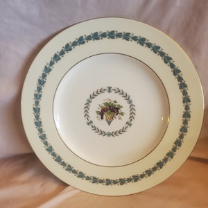 May include: A round, cream-coloured plate with a gold rim and a teal leaf pattern border. The centre features a basket of grapes and leaves. This decorative plate is perfect for serving food.