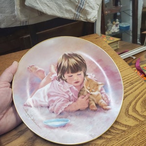 May include: A porcelain plate with a pink background featuring a young girl in a pink dress holding a small orange kitten. The girl is looking down at the kitten and the kitten is looking up at the girl. The plate has a gold rim and the artist's signature is in the bottom left corner.