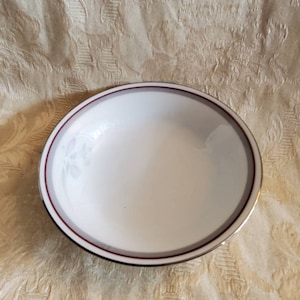 Puede incluir: Un tazón de cerámica blanco con un borde rojo y plateado. El tazón tiene un diseño floral en el interior.