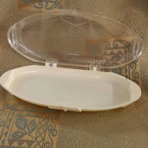May include: A white plastic container with a clear plastic lid. The container is oval shaped and has a smooth surface.