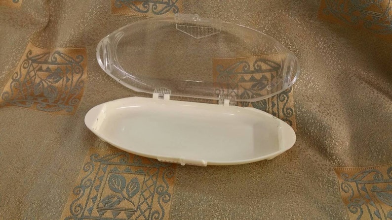 May include: A white plastic container with a clear plastic lid. The container is oval shaped and has a smooth surface.