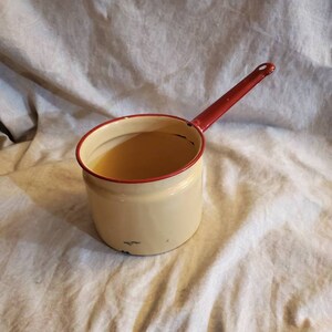 May include: A cream-colored enamel saucepan with a red handle and rim. The saucepan contains a liquid. The handle has a hole at the end for hanging. The saucepan shows signs of wear.