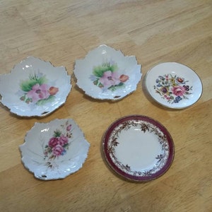 Puede incluir: Cinco platos decorativos de porcelana. Tres tienen forma de hoja con diseños florales en rosa, verde y blanco. Dos son redondos, uno con un estampado floral y otro con un borde burdeos.