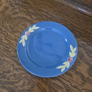 May include: A round, blue ceramic plate with a floral design. The plate features yellow leaves and pink roses on the rim. The plate is sitting on a wooden surface.