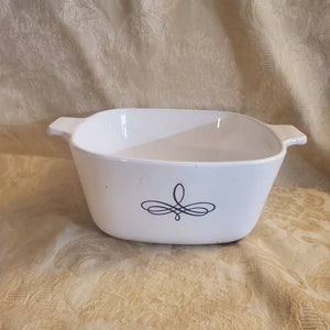 May include: White, square casserole dish with two handles. A black, stylized floral design is centered on the front. The dish is empty and appears to be made of ceramic or a similar material.