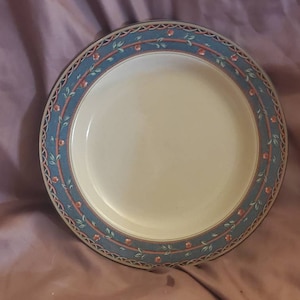 Könnte beinhalten: Ein weißer Keramik-Dinnerteller mit einem blauen und grünen floralen Rand. Der Rand zeigt ein sich wiederholendes Muster aus rosa Blüten und grünen Blättern.