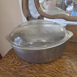 Può includere: Una pirofila vintage in metallo grigio con coperchio in vetro trasparente. Il coperchio presenta un disegno a foglie inciso. La pirofila è su una superficie di legno, con uno specchio decorativo sullo sfondo.