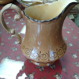 May include: A brown ceramic jug with a decorative swirl design. The jug has a wide mouth and a handle.