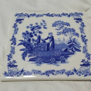 May include: A blue and white ceramic tile with a floral border and a scene of a woman in a garden. The woman is holding a pitcher and standing near a fountain.