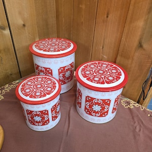 May include: Set of three vintage metal canisters. The canisters are white with red lids and decorative red floral patterns. The canisters are cylindrical and vary in size.
