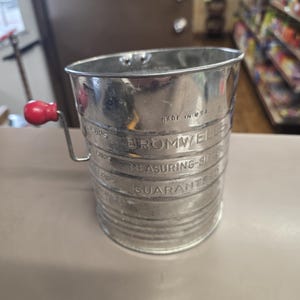 May include: A vintage metal sifter with a red handle. The sifter is cylindrical with the text "BROMWELLS MEASURING-SIFTER GUARANTEED" and "MADE IN USA" printed on the side. It is designed for measuring and sifting ingredients.