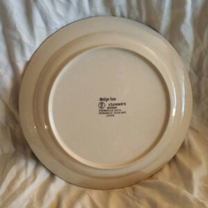 May include: The back of a white ceramic dinner plate with a brown rim. The plate has a brown and white design around the edge. The plate has the words "Design for Stoneware" printed on the bottom.