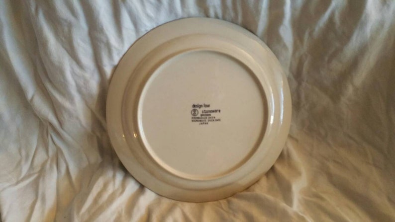 May include: The back of a white ceramic dinner plate with a brown rim. The plate has a brown and white design around the edge. The plate has the words "Design for Stoneware" printed on the bottom.