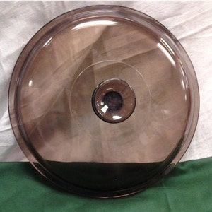 Puede incluir: Una tapa de vidrio transparente con un tinte marrón y una perilla en el centro. La tapa es redonda y tiene una superficie lisa.