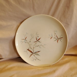 May include: A white ceramic dinner plate with a floral design featuring brown and green leaves and blue flowers.