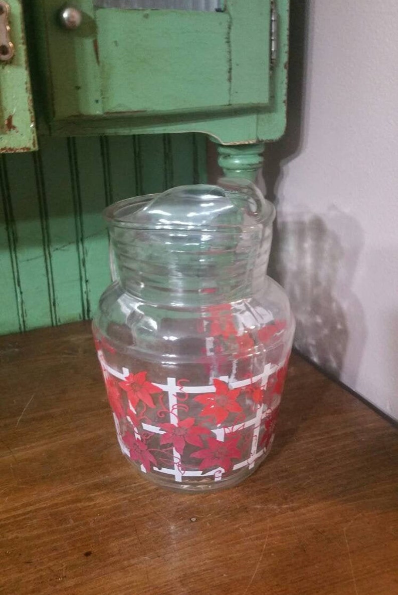 May include: A clear glass pitcher with a red and white floral design. The pitcher has a lid and is perfect for serving beverages.