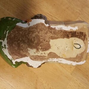 May include: A ceramic wall hanging in the shape of a person with a green hat and red hair. The back of the wall hanging is white with a brown stain and a hole for hanging.