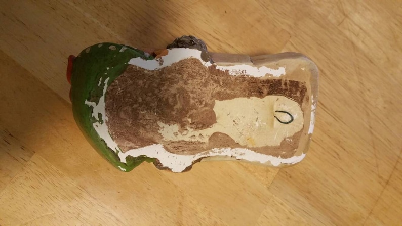 May include: A ceramic wall hanging in the shape of a person with a green hat and red hair. The back of the wall hanging is white with a brown stain and a hole for hanging.