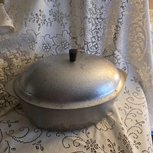 May include: A vintage, silver-colored cooking pot with a lid. The pot has a textured surface and a black knob on top of the lid. The pot has two small handles on either side. The pot is sitting on a white lace tablecloth.