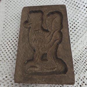 Puede incluir: Un molde de madera rectangular y antiguo con un diseño tallado de un pájaro. El pájaro mira hacia la izquierda con un cuello largo y un cuerpo detallado. La madera es de color marrón oscuro y muestra signos de desgaste.