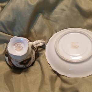 May include: A white teacup and saucer set with a gold and brown floral design. The teacup has a scalloped rim and the saucer has a scalloped edge.
