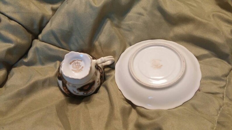 May include: A white teacup and saucer set with a gold and brown floral design. The teacup has a scalloped rim and the saucer has a scalloped edge.