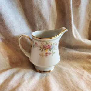 May include: A white porcelain creamer with a floral pattern in pink, blue, and yellow. The creamer has a gold rim and handle, and a pedestal base with a gold band. The creamer is designed for serving cream or milk.