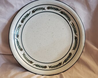 Op de afbeelding: Witte keramische dinerbord met een groen en bruin ontwerp van dennennaalden en kegels rond de rand.
