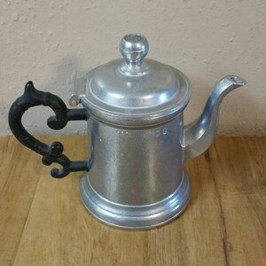 May include: A silver teapot with a black handle and a lid. The teapot has a round body and a spout. The teapot is sitting on a wooden surface.