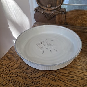 Może przedstawiać: Białe ceramiczne naczynie do ciasta z jasnoniebieskim obramowaniem i wzorem pszenicy pośrodku. Naczynie ma żebrowany brzeg i stoi na drewnianej powierzchni.