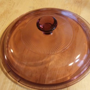 Puede incluir: Una tapa de vidrio transparente con una perilla morada. La tapa tiene un diseño texturizado y acanalado.