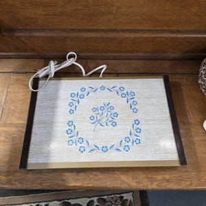 Puede incluir: Bandeja eléctrica rectangular con superficie beige y diseño floral azul. La bandeja tiene un borde marrón y un cable de alimentación blanco. El diseño floral es una corona circular de flores azules.