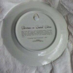 Pu&ograve; includere: Un piatto in ceramica bianca con un bordo dorato e una scritta dorata che recita "Christmas in the United States". La scritta &egrave; circondata da un bordo dorato. Il piatto ha un foro al centro per appenderlo.