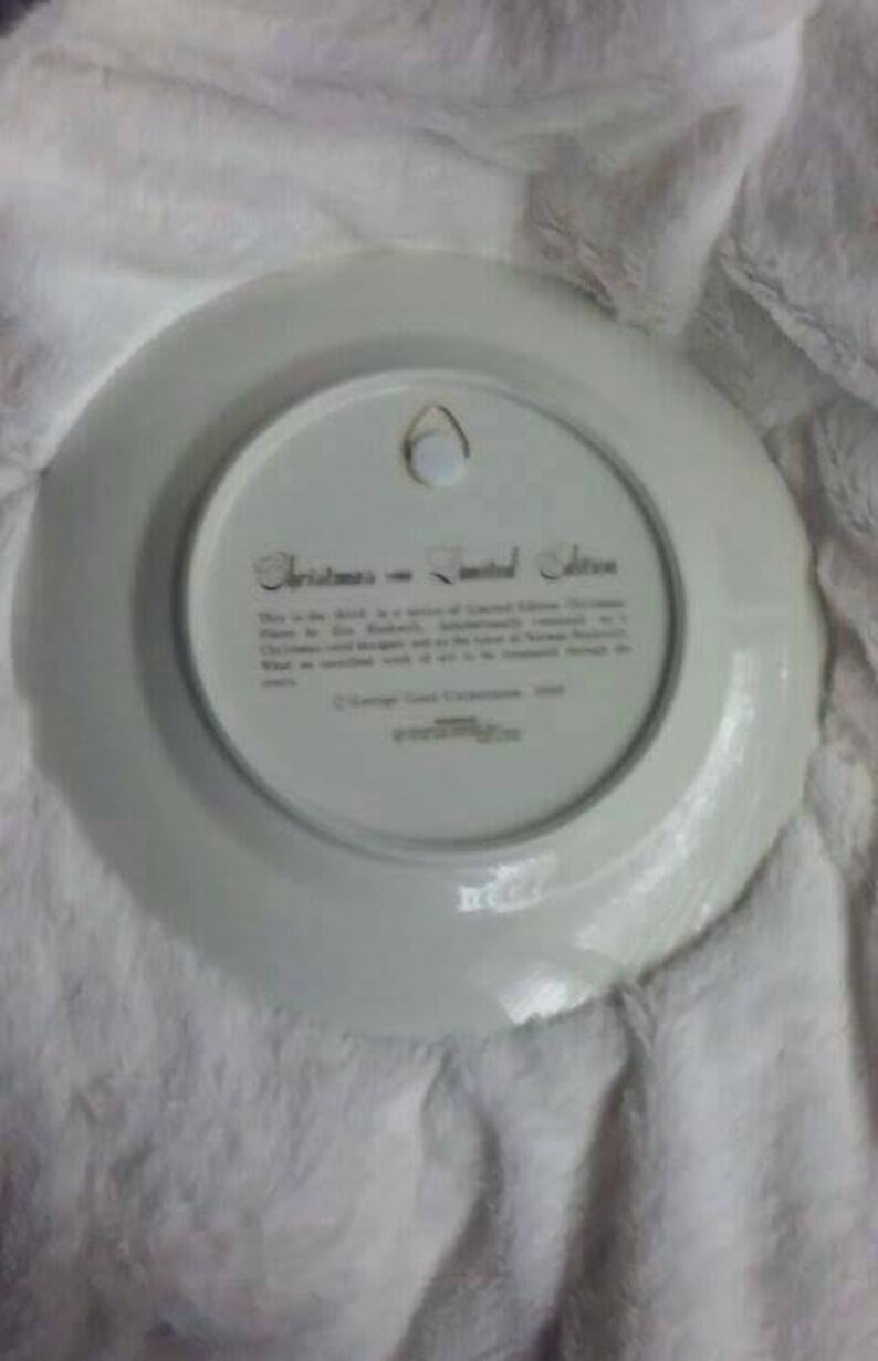 Pu&ograve; includere: Un piatto in ceramica bianca con un bordo dorato e una scritta dorata che recita "Christmas in the United States". La scritta &egrave; circondata da un bordo dorato. Il piatto ha un foro al centro per appenderlo.
